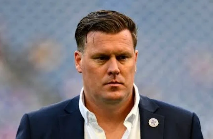 A headshot portrait of football coach Otto Björkegren, who has been appointed to lead the Black Stars interim technical team. He is captured with a serious expression, wearing a navy blue blazer and a white open-collar shirt against a blurred stadium background.