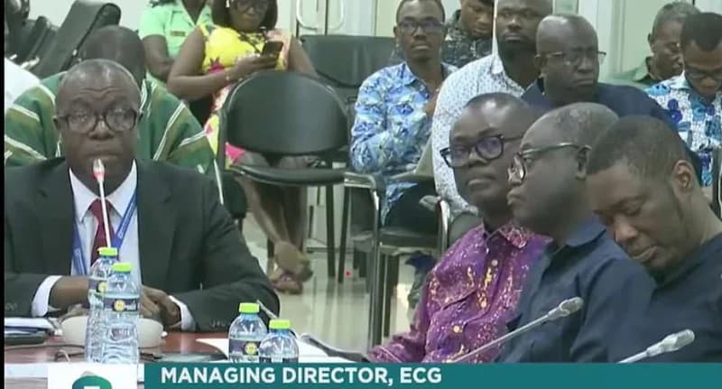 The image depicts the Managing Director of the Electricity Company of Ghana (ECG) appearing before a committee, likely the Public Accounts Committee. He is seen speaking into a microphone at a table with several water bottles, while other officials and observers sit in the background.