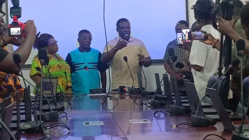 A central government official gestures while speaking at a press conference, surrounded by colleagues and media personnel. The scene is set in a conference room with a long table featuring microphones and laptops, as journalists record the address with cameras and phones.
