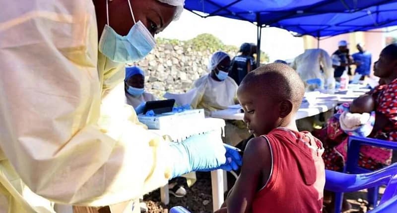 A healthcare worker wearing full personal protective equipment administers a vaccine to a young child at an outdoor clinic. The setting features blue canopies and chairs, with other medical staff and community members visible in the background, illustrating a public health campaign.