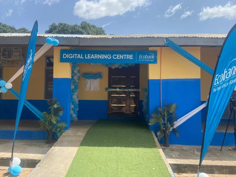 The image shows the entrance of a newly established Digital Learning Centre commissioned by Ecobank. The building is painted in bright blue and yellow, decorated with blue and white balloons and brand banners, featuring a green artificial turf walkway leading to the ribbon-adorned entrance.