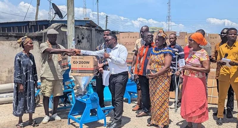 Officials from the Cape Coast Metropolitan Assembly (CCMA) are pictured during a presentation ceremony where they are donating industrial equipment to Persons With Disabilities. A man in a white shirt and black trousers is seen handing over a blue grinding machine to a recipient, while other community members and officials observe the donation.