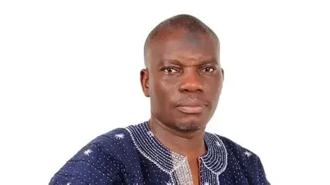 A professional portrait of Mustapha Gbande, a Deputy General Secretary of the National Democratic Congress (NDC), looking directly at the camera. He is wearing a traditional blue and white patterned smock, a common attire for political and social events in Ghana.