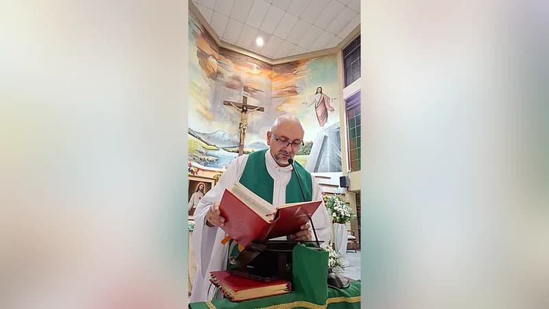 A priest dressed in a white robe and green stole is seen reading from a large red liturgical book at a lectern. Behind him is a large church mural depicting religious scenes, including a prominent crucifix mounted on the wall.