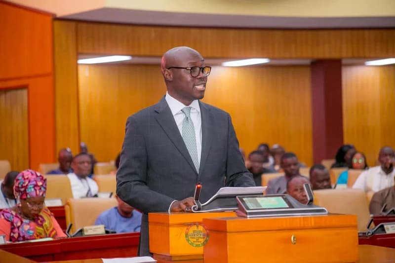 Dr. Cassiel Ato Forson, a prominent Ghanaian politician, is seen speaking at a podium inside the Parliament House in Accra. He is dressed in a formal suit and glasses, addressing the chamber during a session, with other members of parliament visible in the background.