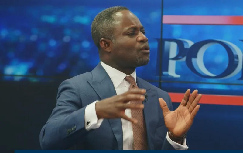 Kwabena Mintah Akandoh, a Ghanaian Member of Parliament, is pictured in a television studio making a point with expressive hand gestures. He is dressed in a professional blue suit and patterned tie against a digital studio backdrop with a news program graphic.