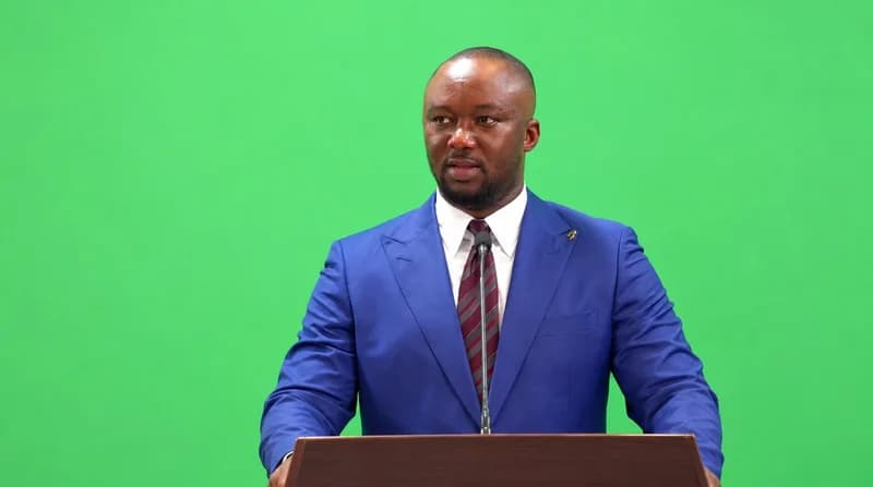A man, likely the CEO of the Youth Employment Agency (YEA), Kofi Baah Agyepong, is captured during a presentation. He stands behind a wooden podium with a microphone, wearing a blue suit and a striped tie against a solid green background.