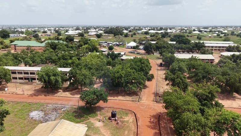 An aerial view of the Bawku Senior High School campus in Ghana, showing several school buildings interspersed with lush green trees. Red dirt paths connect different parts of the facility, and students in pink uniforms are seen near a water storage tank.