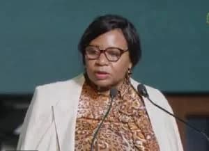 A woman with glasses and a white blazer over a leopard-print blouse is captured speaking at a formal event. She is positioned behind a pair of microphones against a dark teal background, likely delivering a speech or presentation related to African economic affairs.