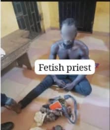 A man identified as a fetish priest sits shirtless on a tiled floor with various ritual items placed in front of him. The subject's face is blurred, and a text box reading "Fetish priest" is overlaid on the image, which appears to be taken in a detention or interrogation setting.