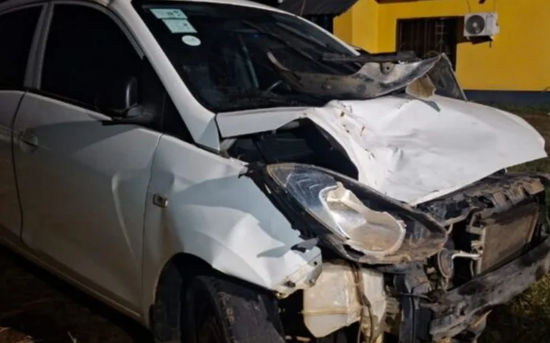 A white vehicle exhibits severe front-end damage with a heavily crumpled hood and exposed internal parts. The car is positioned outdoors in front of a bright yellow building, showing the aftermath of a significant impact.
