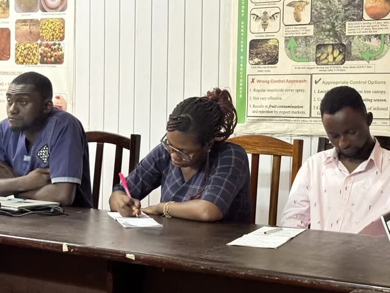 Three academics are seated at a long wooden table during a training session or lecture at the University of Ghana. A woman in the center is diligently taking notes, while colleagues sit beside her in front of a wall featuring scientific posters related to agricultural pest control and regulatory services.