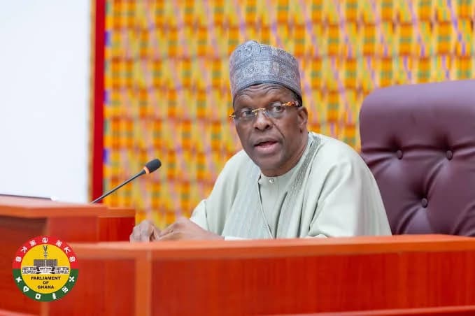 Alban Bagbin, the Speaker of the Parliament of Ghana, is pictured at the speaker's dais during a parliamentary session. He is dressed in traditional Ghanaian attire and a cap, with the official seal of Parliament visible in the bottom-left corner.