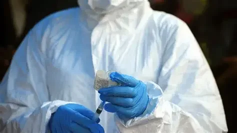 A medical worker wearing a white full-body personal protective equipment (PPE) suit and blue surgical gloves is shown preparing a medical vial and syringe. The image captures the intensive safety measures and clinical environment associated with infectious disease management.