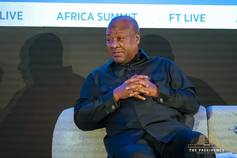 President John Dramani Mahama is pictured seated during a high-level discussion at an international forum. He is wearing a black traditional-style shirt with his hands clasped, appearing in a contemplative pose against a backdrop featuring 'Africa Summit' branding.