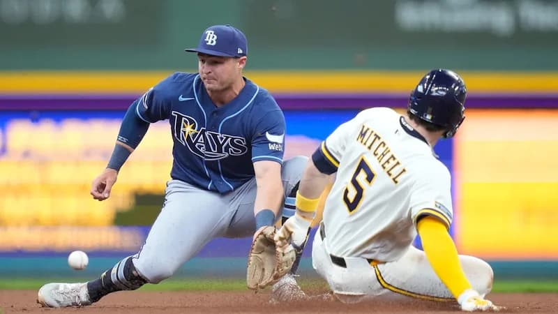 A high-action photograph showing Milwaukee Brewers player Garrett Mitchell (number 5) sliding into a base as a Tampa Bay Rays fielder prepares to catch a bouncing baseball. The scene captures the dynamic energy and intense competition of a Major League Baseball game between these two franchises.