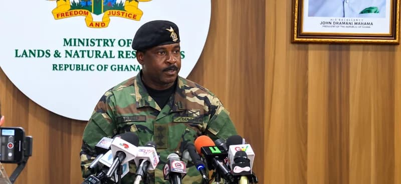 A Ghanaian military officer in camouflage uniform and a black beret addresses the press at the Ministry of Lands and Natural Resources. He is positioned behind multiple microphones, with the ministry's seal and a portrait of President John Dramani Mahama visible in the background.
