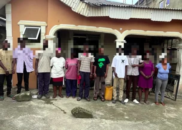 A photograph showing eleven individuals standing in a row in front of a residential building, likely the 11 Ghanaians rescued from a visa scam in Nigeria. Their faces are blurred to protect their identities, and they are standing on a concrete patio area with some appearing to have their hands behind their backs.