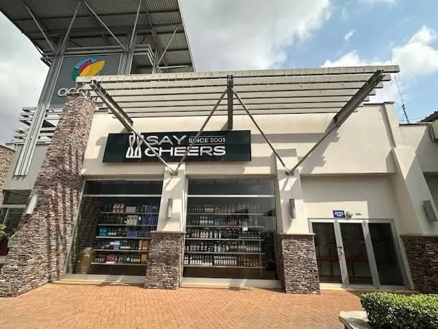 The photograph shows the exterior of a Say Cheers retail outlet, a prominent liquor store located in a modern commercial complex. The storefront features large glass windows displaying neatly arranged bottles on shelves, framed by rustic stone pillars and a large black sign with white lettering.