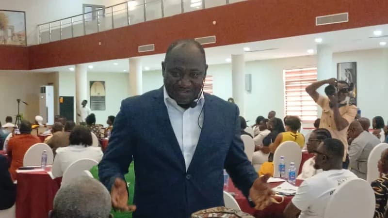 A man in a dark blazer addresses an audience at a formal gathering in a large hall, likely related to the Sunyani Traditional Council's development agenda launch. Attendees are seated at tables with red cloths, and a photographer is seen in the background recording the proceedings.