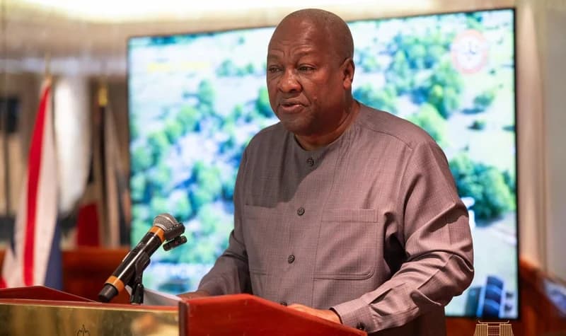 President John Mahama is captured speaking at a lectern during a high-level meeting in London. He is wearing a grey traditional-style shirt and standing before a microphone, with a large digital screen behind him displaying an aerial green landscape.