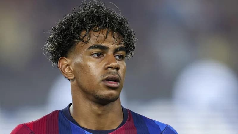 A close-up portrait of professional footballer Lamine Yamal wearing a blue and red striped FC Barcelona jersey. The young athlete has dark curly hair and a serious expression, looking slightly off-camera against a blurred stadium-like background.