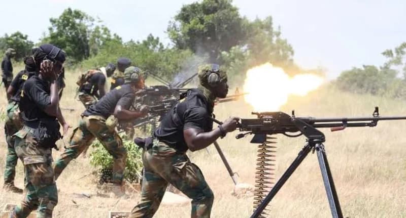 Soldiers from the Ghana Armed Forces engage in a live-fire training exercise at a range. A soldier in the foreground is captured at the moment of firing a heavy machine gun, producing a large muzzle flash, while colleagues in tactical gear observe and manage equipment in the background.