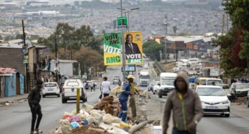 A busy South African street scene featuring large African National Congress (ANC) campaign posters with the image of Cyril Ramaphosa and the slogan 'VOTE ANC'. In the foreground, piles of waste sit on the road median as pedestrians and vehicles navigate the congested thoroughfare. The image highlights the juxtaposition of political messaging against the backdrop of infrastructure and service delivery challenges.