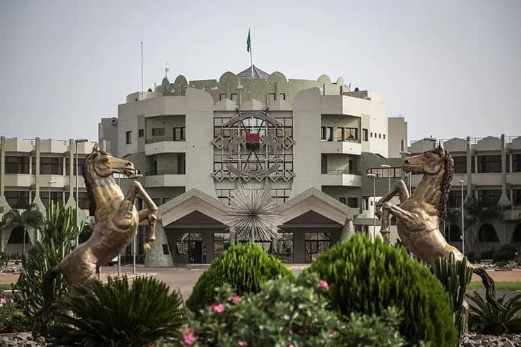 A photograph of the National Assembly building in Ouagadougou, Burkina Faso, featuring its distinctive architecture and central state emblem. In the foreground, two large bronze-colored statues of rearing horses stand amidst manicured green shrubbery and pink flowers under a clear sky.