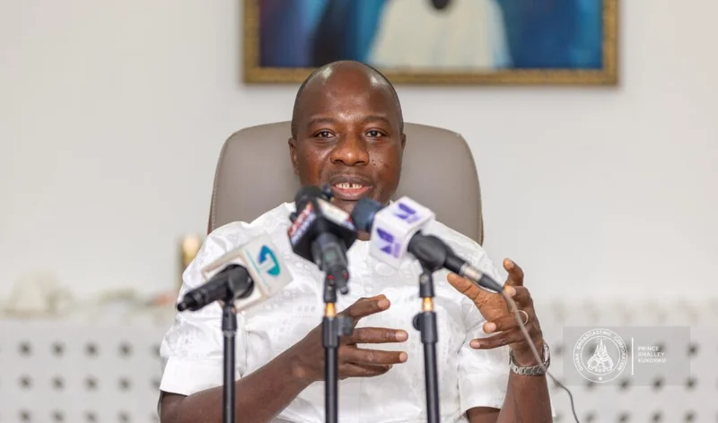 Mahama Ayariga, the Member of Parliament for Bawku Central, is seen speaking at a press engagement. He is seated behind several microphones from different news organizations, using hand gestures to emphasize his points.