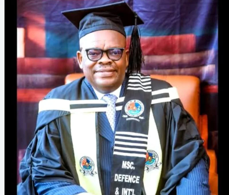 A formal portrait of celebrated Ghanaian actor and TV personality Kwame Dzokoto wearing academic regalia. He is dressed in a black graduation gown and mortarboard, with a stole that identifies him as a graduate of a Master of Science program in Defence and International Affairs.