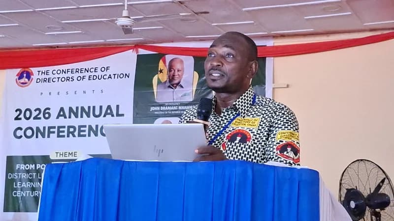 A speaker addresses the 2026 Annual Conference of the Conference of Directors of Education (CODE) in Ghana. The man stands at a blue-draped podium with a laptop, set against a backdrop featuring a portrait of President John Dramani Mahama.