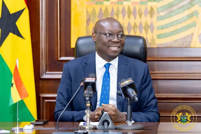 Dr. Mohammed Amin Adam, Ghana's Finance Minister, addresses the media during a press briefing. He is seated at a desk featuring the national flag, several media microphones, and the official Ministry of Finance seal in the background.