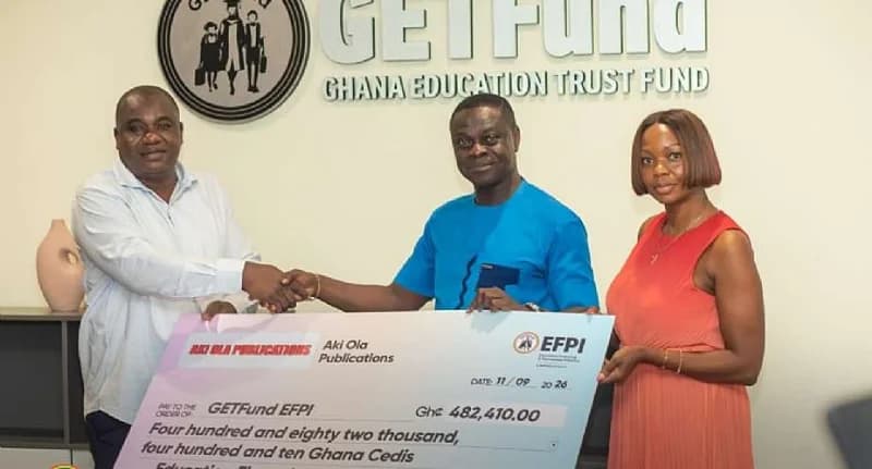 A formal presentation ceremony where representatives from Aki-Ola Publications donate a symbolic check worth GH¢482,410 to the Ghana Education Trust Fund (GETFund). Two men shake hands behind the large check while a woman in a red dress assists in holding the document, all standing in front of a GETFund logo on an office wall.