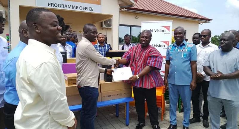 Representatives from World Vision Ghana are seen handing over IT equipment and learning materials to local officials in Agotime-Ziope. The central figures are shaking hands over a boxed laptop in front of a 'Programme Office' building, with stacks of equipment visible on a blue table. The scene captures a formal donation event aimed at supporting education in the region.