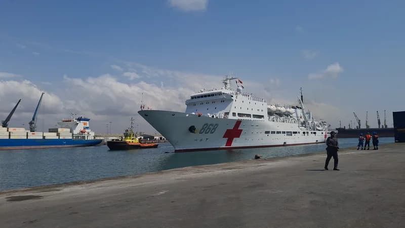 A large white Chinese hospital ship with the hull number 866 and a prominent red cross symbol arrives at a port, escorted by a small tugboat. Personnel and onlookers are visible on the concrete pier as the vessel prepares to dock under a bright, partly cloudy sky.