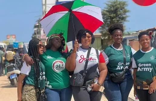 A group of five women, supporters of the National Democratic Congress (NDC), pose together outdoors under a party-branded umbrella. They are wearing campaign apparel featuring the name of John Mahama and the 'Green Army' slogan, indicating active political participation in Ghana.