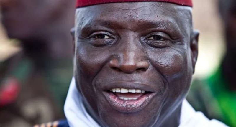 A close-up portrait of former Gambian President Yahya Jammeh, wearing his signature red kufi hat and white traditional attire. He is captured with a slight smile and direct gaze towards the camera.