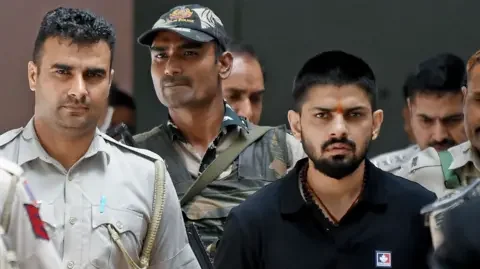 Reuters Policemen escort Lawrence Bishnoi at a court, in New Delhi, India on 18 April 2023. He is wearing a black t-shirt and has a tilak (saffron mark) on forehead.