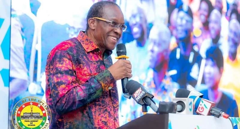 Alban Bagbin, the Speaker of the Parliament of Ghana, is pictured smiling while speaking at a podium during a public event. He is wearing a vibrant, multicolored patterned shirt and holding a microphone, with several media microphones visible in the foreground including Joy News and GTV.