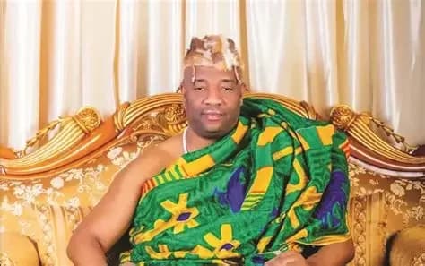 A portrait of King Tackie Teiko Tsuru II, the Ga Mantse, dressed in traditional Ghanaian Kente cloth and animal skin headgear. He is seated on an ornate gold throne against a backdrop of light-colored curtains.