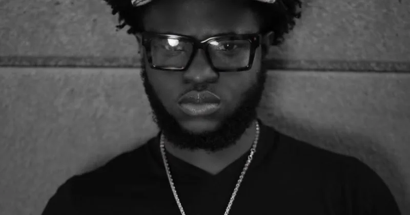 A close-up black and white portrait of the Ghanaian emerging musician BLACQ GOAT. He is shown wearing thick-rimmed glasses, a silver-toned chain, and a cap while looking directly at the camera with a serious expression.