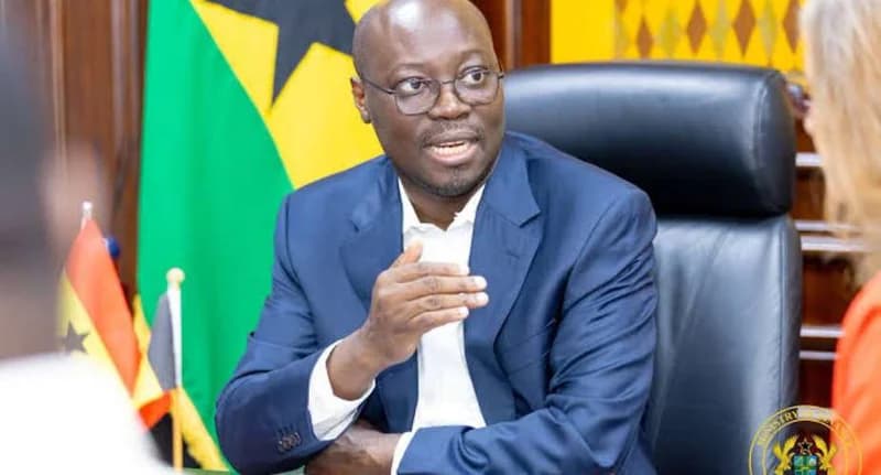 Dr. Cassiel Ato Forson, a prominent Ghanaian politician, is seen gesturing while speaking in an official setting. He is seated in a large black leather chair with the national flag of Ghana visible in the background, suggesting a formal government or parliamentary context.