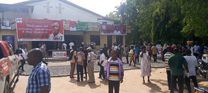 A crowd of people gathers outside a building decorated with National Democratic Congress (NDC) campaign banners for a conference or election event. The scene features prominent posters for candidates such as Hayford Amanowu and Fred Kwasi Agbedi, with a visible police checkpoint barricade suggesting a secured political function.