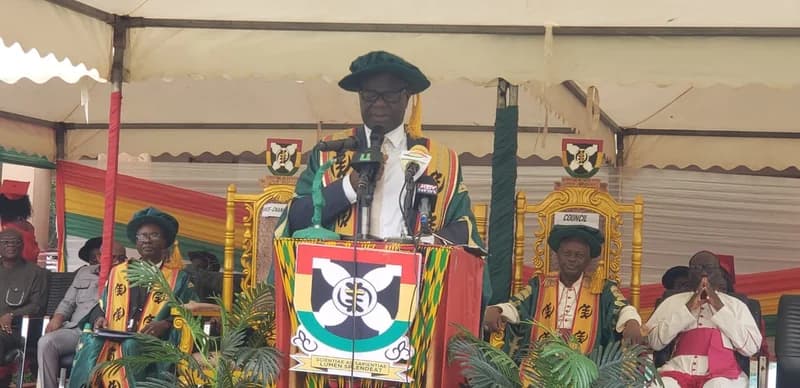 An official in green and gold academic regalia delivers a speech from a podium during a graduation ceremony at the Catholic University of Ghana. He is flanked by other university officials and religious leaders seated under a white canopy decorated with Ghanaian national colors.