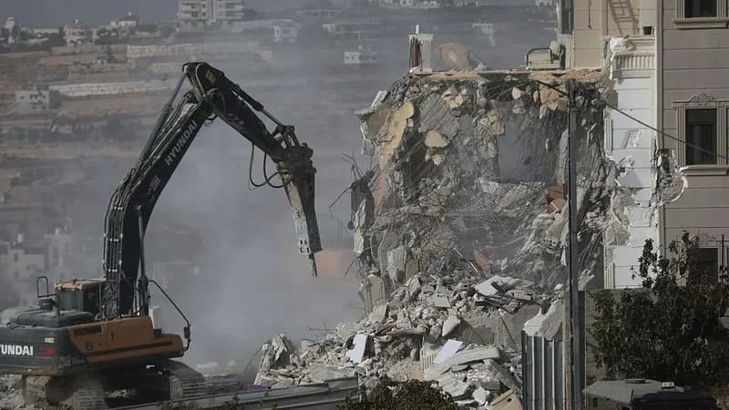 A large Hyundai excavator equipped with a hydraulic breaker is seen demolishing a multi-story residential building. Dust and debris fill the air as the structure is reduced to rubble, with exposed steel reinforcement visible in the wreckage.