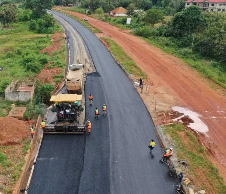 An aerial photograph capturing the ongoing asphaltic works on the Sunyani inner ring road project. A heavy-duty yellow paver and a dump truck are shown laying a fresh layer of dark asphalt while several construction workers in high-visibility gear monitor the progress along the road corridor.