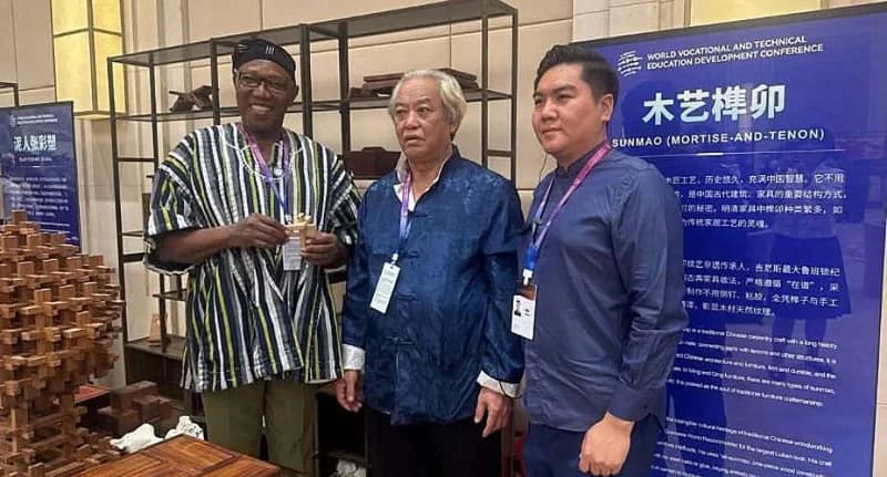 A Ghanaian representative wearing a traditional fugu smock stands alongside two Chinese officials at the World Vocational and Technical Education Development Conference. They are positioned in front of an educational display showcasing 'Sunmao' (mortise-and-tenon) carpentry techniques, symbolizing the technical exchange between the two nations.