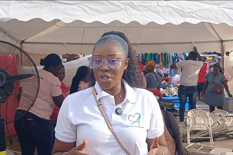 Ghanaian media personality Frema Adunyame is seen speaking to a camera under a white event canopy. She is wearing blue-rimmed glasses and a white polo shirt featuring a logo for her foundation, with a busy background showing event preparations including chairs, a fan, and other attendees.