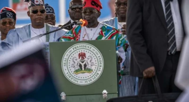 Nigerian President Bola Ahmed Tinubu is pictured speaking at a podium featuring the official Seal of the President. He is wearing a red cap and traditional attire with colors associated with the All Progressives Congress (APC) party, surrounded by several men in a political or official setting.